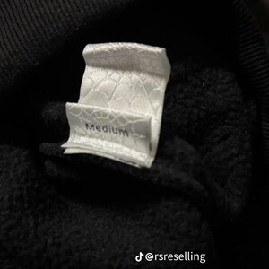 Puede incluir: Primer plano de una etiqueta de ropa blanca con la palabra "Medium" impresa. La etiqueta tiene un patr&oacute;n texturizado y est&aacute; adherida a una tela negra. Probablemente sea una prenda de vestir.