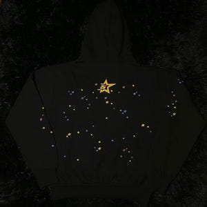 May include: Black hooded sweatshirt with a star design. The back of the sweatshirt features a gold star graphic and scattered smaller stars in yellow, blue, and purple. The sweatshirt has a drawstring hood and a kangaroo pocket.