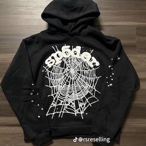 Puede incluir: Sudadera con capucha negra con un dise&ntilde;o de telara&ntilde;a blanca y la palabra "sp5der" en una fuente estilizada. Acentos de estrellas blancas en las mangas. La sudadera tiene un bolsillo delantero y una capucha con cord&oacute;n.