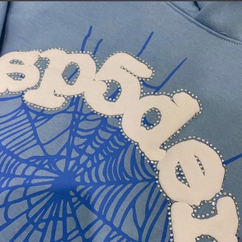 May include: Light blue hoodie with a white and silver rhinestone embellished design. The design features the word "SPIDER" in large, stylized letters, with a blue spiderweb graphic underneath. The hoodie is made of a soft material.