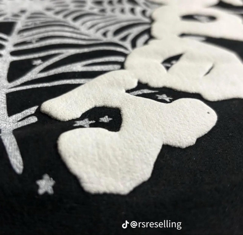 Puede incluir: Primer plano de un textil negro con un dise&ntilde;o blanco en relieve que presenta una calavera estilizada y una telara&ntilde;a. El dise&ntilde;o incluye estrellas y un elemento similar a un &aacute;rbol. El identificador de Instagram @rsreselling es visible.