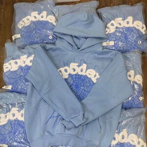 May include: Light blue hoodies with white text and a blue spiderweb design. The word "p5de" is printed across the chest. Several hoodies are visible, some in plastic packaging. The hoodies have a hood and a front pocket.