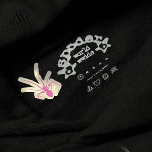 May include: Close-up of a black hoodie with a white and pink spider graphic and the words "world wide" in a circular design. The hoodie also features care symbols and a tag.