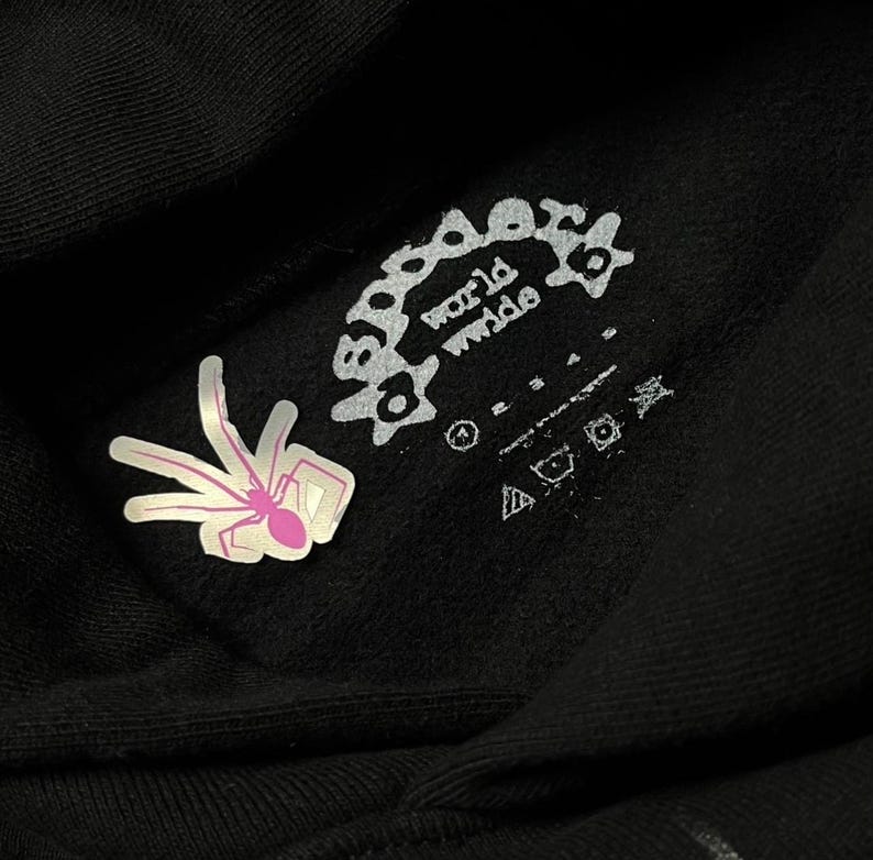 May include: Close-up of a black hoodie with a white and pink spider graphic and the words "world wide" in a circular design. The hoodie also features care symbols and a tag.
