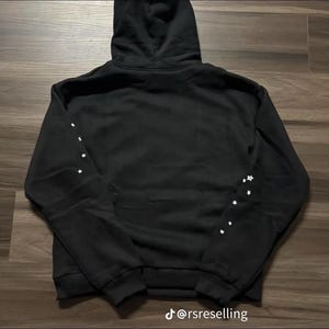Puede incluir: Sudadera con capucha negra con detalles de estrellas blancas en las mangas. La sudadera tiene una capucha con cord&oacute;n y pu&ntilde;os y dobladillo de canal&eacute;. El texto "@rsreselling" est&aacute; en la parte inferior de la imagen.