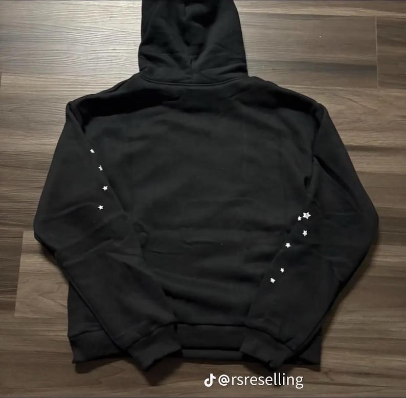 Puede incluir: Sudadera con capucha negra con detalles de estrellas blancas en las mangas. La sudadera tiene una capucha con cord&oacute;n y pu&ntilde;os y dobladillo de canal&eacute;. El texto "@rsreselling" est&aacute; en la parte inferior de la imagen.
