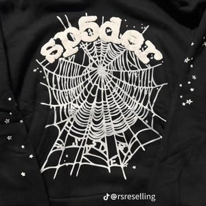 Puede incluir: Sudadera negra con un dise&ntilde;o de telara&ntilde;a blanca y el texto "sp5der" en la parte superior. Peque&ntilde;as estrellas blancas adornan la prenda. El identificador de Instagram "@rsreselling" es visible en la parte inferior.