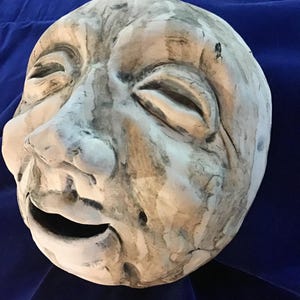 May include: A carved, off-white mask with closed eyes and an open mouth. The mask has a weathered appearance with gray and brown streaks, suggesting age or artistic detail. The background is a deep blue fabric.