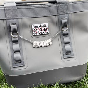May include: A gray tote bag with a baseball mom theme. The bag has a rectangular shape with a darker gray base. It features a patch that says "baseball MOM" and a chain with the name "Sanders". The bag is on green grass.