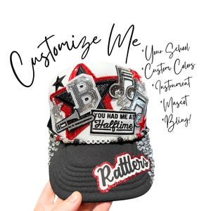 May include: A white and black trucker hat featuring a large silver sequined letter B, musical note patches, and a trombone patch. The text reads "YOU HAD ME AT HALFTIME", "MOMMA", and "Rattlers". The hat is decorated with red and black stars and rhinestones.