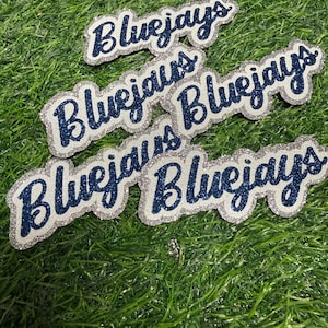 May include: Five individual die-cut labels featuring the word Bluejays written in a cursive script. Each label consists of dark blue glittery lettering layered over a silver glittery background, creating a distinct outline effect for each piece.
