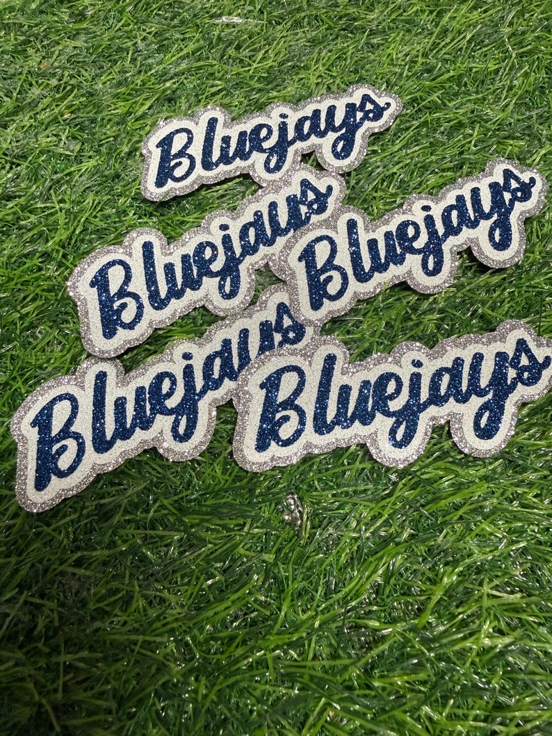 May include: Five individual die-cut labels featuring the word Bluejays written in a cursive script. Each label consists of dark blue glittery lettering layered over a silver glittery background, creating a distinct outline effect for each piece.