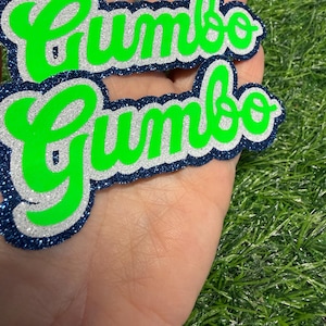 May include: Two layered die-cut signs featuring the word Gumbo in a bright neon green script font. Each sign has a silver glitter middle layer and a dark navy blue glitter outer border, creating a stacked effect with a bold, high-contrast appearance.
