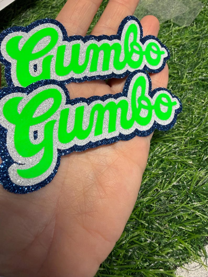 May include: Two layered die-cut signs featuring the word Gumbo in a bright neon green script font. Each sign has a silver glitter middle layer and a dark navy blue glitter outer border, creating a stacked effect with a bold, high-contrast appearance.