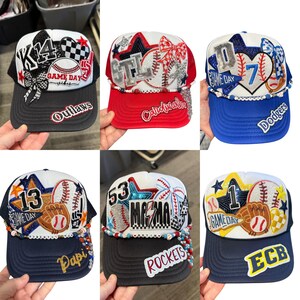 May include: A selection of baseball-themed trucker caps. Each cap has a unique design with text, numbers, and baseball-related embellishments. Colours include black, red, blue, and white. The caps have curved peaks and mesh backs.