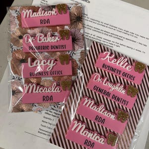 May include: Pink and white personalized name tags with gold glitter tooth accents. The tags include names and titles such as "RDA", "Pediatric Dentist", and "Business Office". The tags are packaged on floral and striped backgrounds.