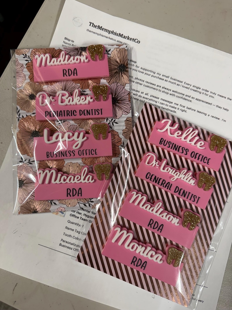 May include: Pink and white personalized name tags with gold glitter tooth accents. The tags include names and titles such as "RDA", "Pediatric Dentist", and "Business Office". The tags are packaged on floral and striped backgrounds.