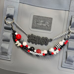 May include: A beaded keychain with a baseball theme. The keychain features a black nameplate that says "Jones" and a beaded strand with baseballs, red, black, and white beads, and the word "GAMEDAY". The keychain is attached to a gray bag.