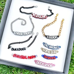 May include: A collection of personalized bracelets with various chain styles and charms. The charms include names like "Katelyn", "Cheer Mom", "Colton", and "Bulldogs". Other charms include "Go Braves", "Game Day", "Dragons", "Baseball Mom", and "Bridesmaid".