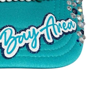 May include: A teal baseball cap featuring the text Bay Area in a white and dark blue glitter script font. The brim is decorated with clear and dark blue rhinestones along the side, and a row of white beads lines the top edge of the cap.