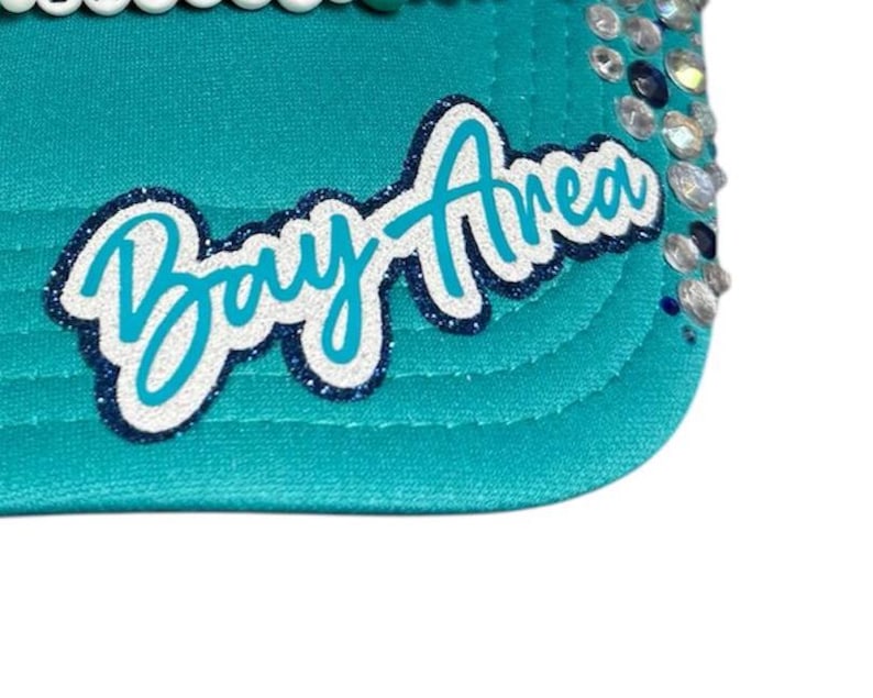 May include: A teal baseball cap featuring the text Bay Area in a white and dark blue glitter script font. The brim is decorated with clear and dark blue rhinestones along the side, and a row of white beads lines the top edge of the cap.