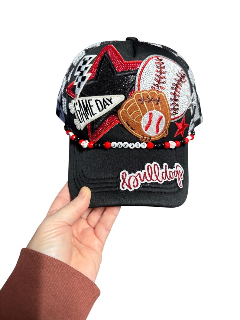 May include: Black baseball cap with a mesh back, featuring a star, baseball, and glove design. The cap has the text "GAME DAY" and "&bulldogs". A beaded band spells out "JAXSON".
