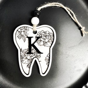 以下が含まれることがあります： 白い木製の歯のオーナメントで、花柄と黒い「K」の文字が描かれています。オーナメントは、黒と白のビーズが付いた紐で吊り下げられています。