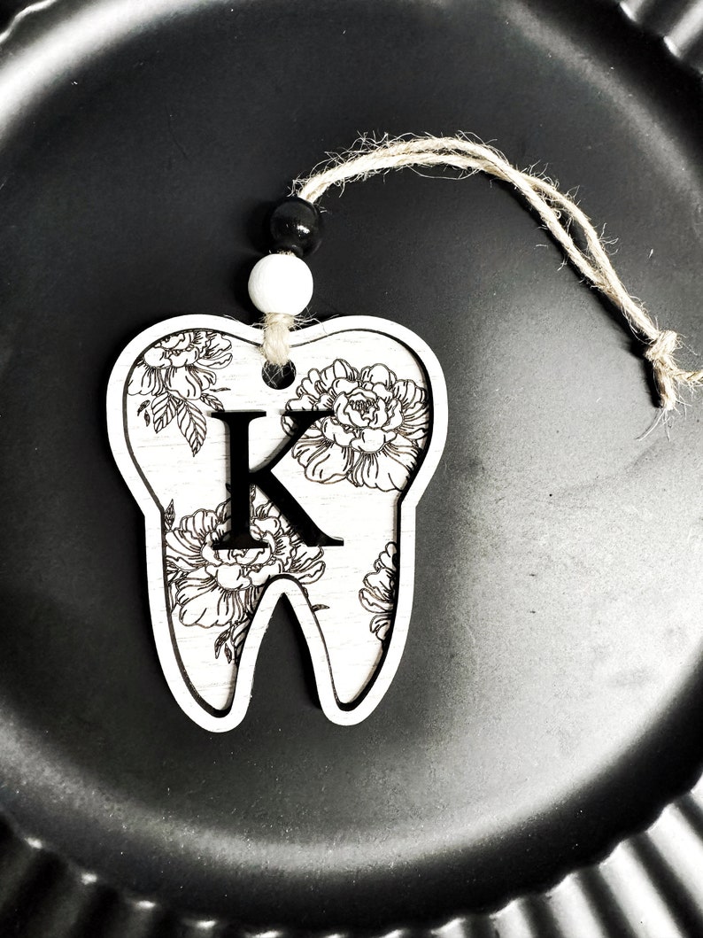 以下が含まれることがあります： 白い木製の歯のオーナメントで、花柄と黒い「K」の文字が描かれています。オーナメントは、黒と白のビーズが付いた紐で吊り下げられています。