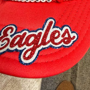 May include: A bright red baseball cap featuring the word Eagles in a glittery red script font with a white and dark blue outline. A row of white beads with black letters is visible along the top edge of the cap brim, adding a decorative detail.