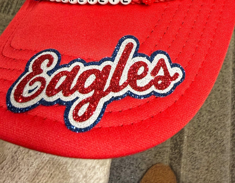 May include: A bright red baseball cap featuring the word Eagles in a glittery red script font with a white and dark blue outline. A row of white beads with black letters is visible along the top edge of the cap brim, adding a decorative detail.