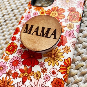 May include: A white phone case with a floral pattern in shades of orange, yellow, pink, and red. A wooden phone grip with the word "MAMA" is attached to the back of the case.
