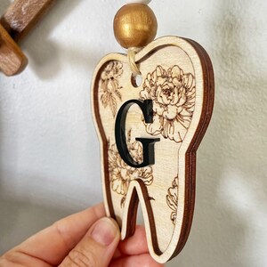 以下が含まれることがあります： 花柄と黒い「G」の文字が書かれた木製の歯の形をしたオーナメント。白いビーズと金色のビーズがついた紐で吊り下げられています。