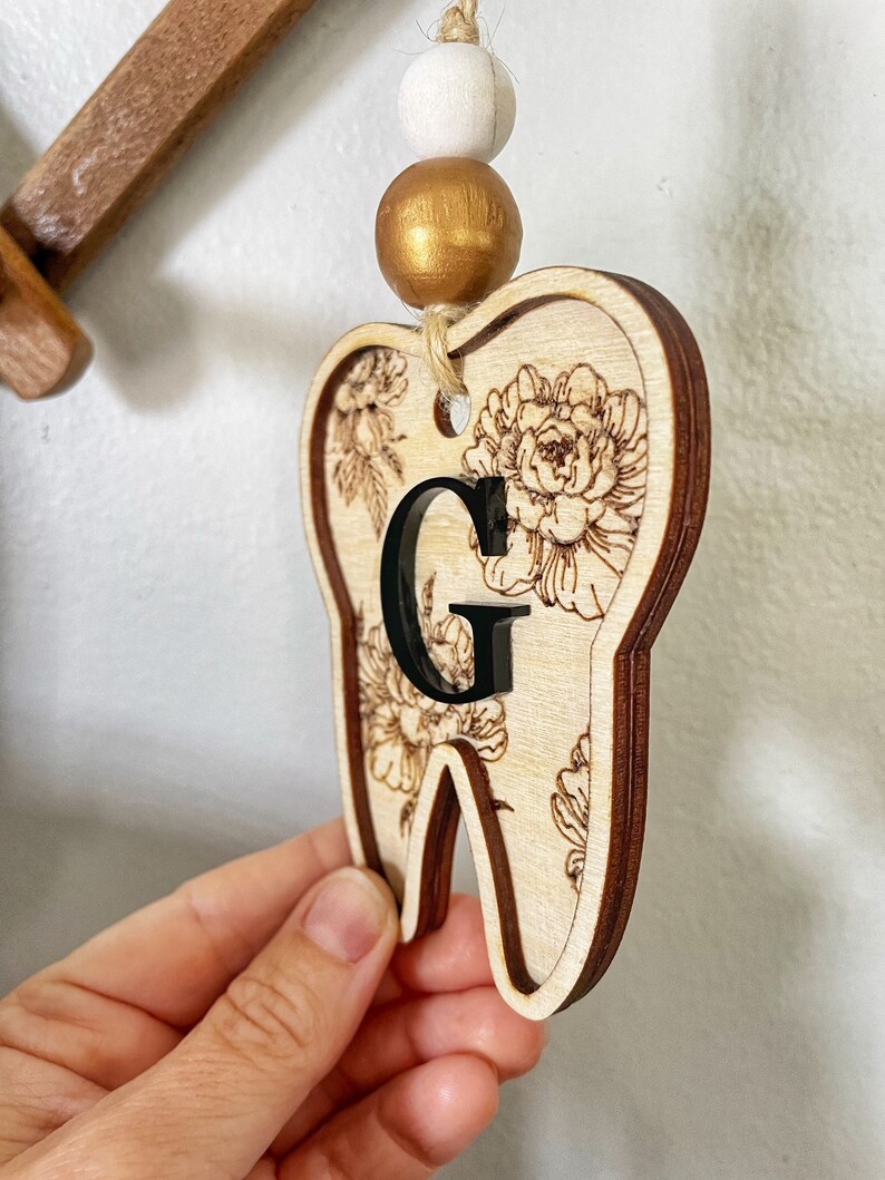 以下が含まれることがあります： 花柄と黒い「G」の文字が書かれた木製の歯の形をしたオーナメント。白いビーズと金色のビーズがついた紐で吊り下げられています。