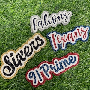 May include: Four glittery, layered die-cut labels featuring the text Falcons in black, Sixers in black, Texans in red, and 91 Prime in blue. Each label has a distinct glitter border in silver, gold, blue, or red, creating a textured, dimensional appearance.
