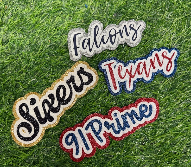 May include: Four glittery, layered die-cut labels featuring the text Falcons in black, Sixers in black, Texans in red, and 91 Prime in blue. Each label has a distinct glitter border in silver, gold, blue, or red, creating a textured, dimensional appearance.