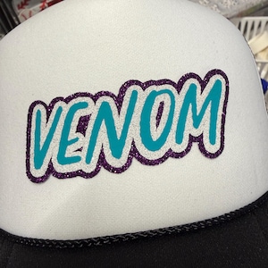 May include: A white trucker hat featuring the word VENOM in teal letters with a silver glitter outline and a dark purple glitter border. The hat has a black brim and a black braided cord detail across the base of the white foam front panel.
