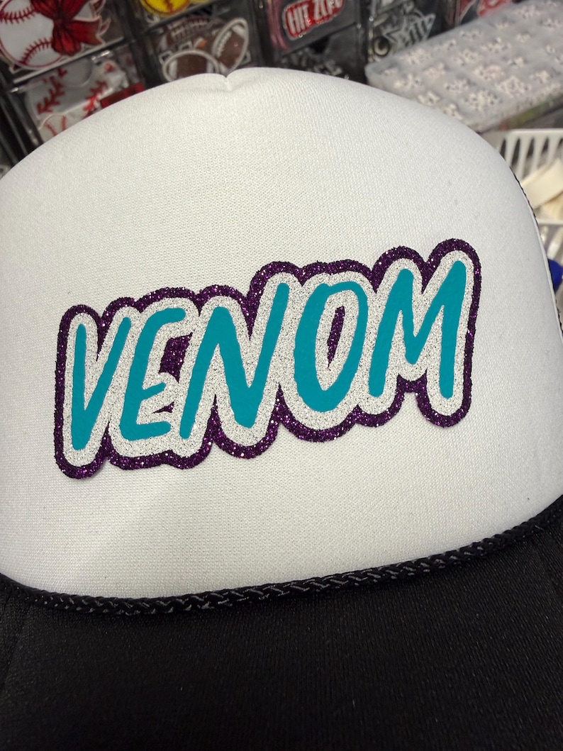 May include: A white trucker hat featuring the word VENOM in teal letters with a silver glitter outline and a dark purple glitter border. The hat has a black brim and a black braided cord detail across the base of the white foam front panel.