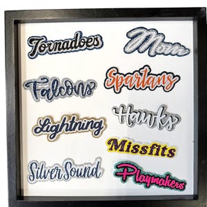 May include: A black square frame containing eight glittery fabric patches with text. The patches read Tornadoes, Mom, Falcons, Spartans, Lightning, Hawks, Missfits, and Playmakers in various colors including silver, gold, orange, purple, and pink.