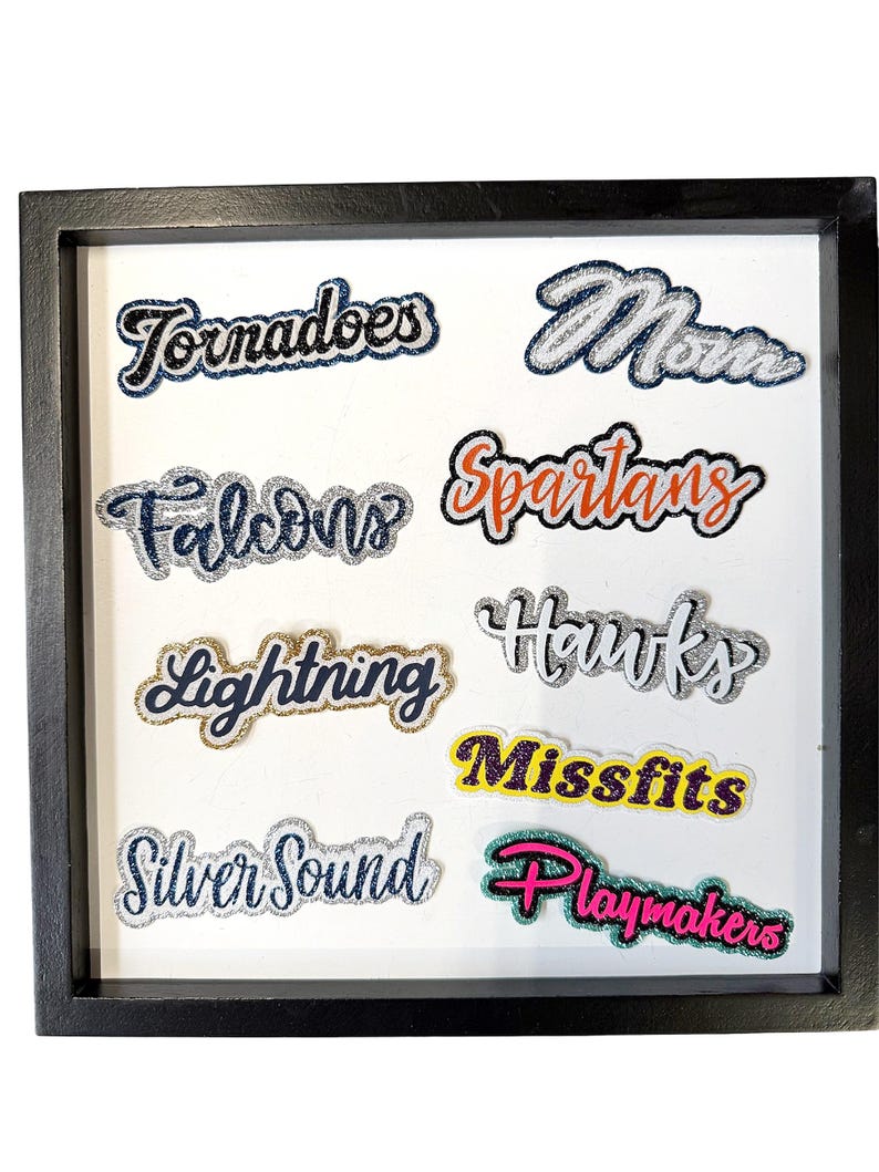 May include: A black square frame containing eight glittery fabric patches with text. The patches read Tornadoes, Mom, Falcons, Spartans, Lightning, Hawks, Missfits, and Playmakers in various colors including silver, gold, orange, purple, and pink.