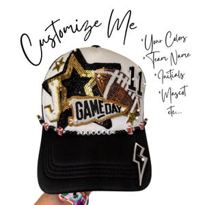 May include: A black and white baseball cap with gold and black embellishments, including a football and the word "GAMEDAY". The cap features the word "ZEBRAS" and the text "Customize Me".