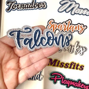 May include: A collection of glittery team name decals, including "Falcons," "Tornadoes," and "Playmakers." The decals feature various color combinations, such as blue, silver, and pink, with a black border. The decals are displayed on a white background.