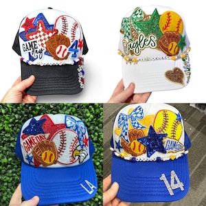 May include: Four baseball caps with decorative embellishments. Each cap features a baseball or softball theme, with elements like baseballs, gloves, stars, and the text "Game Day" or "Eagles". The caps have different colour schemes, including black, white, and blue.