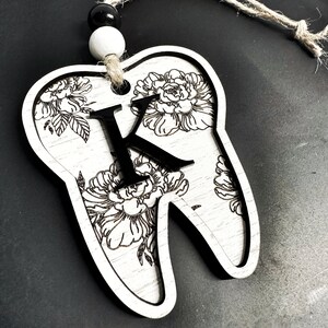 以下が含まれることがあります： 白い木製の歯のオーナメントで、黒い花の模様と中央に「K」の文字があります。オーナメントには、吊り下げ用の麻ひもが付いています。