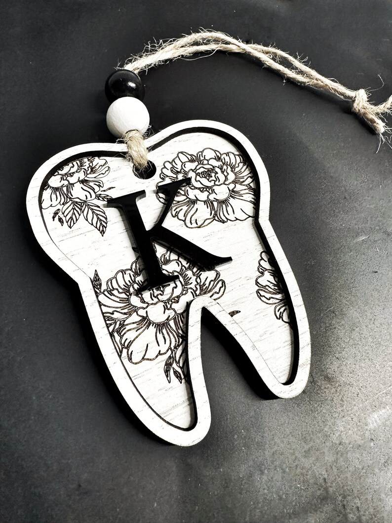 以下が含まれることがあります： 白い木製の歯のオーナメントで、黒い花の模様と中央に「K」の文字があります。オーナメントには、吊り下げ用の麻ひもが付いています。