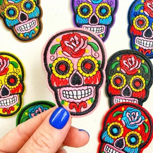 May include: Colorful embroidered skull patches with floral and geometric designs. The patches feature a variety of colors, including pink, red, yellow, blue, and purple. The patches are likely for clothing or accessories.