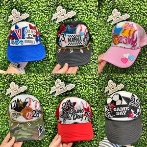 May include: A selection of baseball caps with diverse designs and text. One cap is blue with "EAT. MY. BUBBLES" text. Others feature numbers, stars, baseballs, and the words "MAMA" and "GAME DAY".