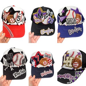 May include: A selection of baseball caps with baseball-themed designs. Each cap features a baseball, star, and team name. Colours include red, black, white, and blue. Some caps have the text "Game Day", "Mama", "Eagles", "Rockies", "Wow Factor", and "Naturals".