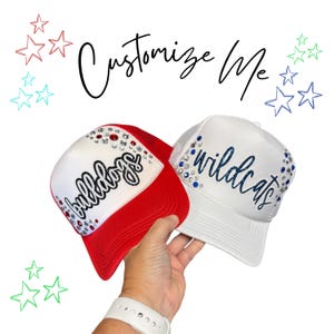 May include: Two trucker hats with mesh backs, one red and white with the word Bulldogs in black script, the other all white with the word wildcats in blue script. Both hats feature rhinestone embellishments and the text Customize Me above.