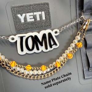 May include: A custom nameplate necklace with the name "TOMA" in black lettering on a white background. The necklace features a chain with faux pearls and small yellow and red softball charms. The text "Name Plate Chain sold separately" is also visible.