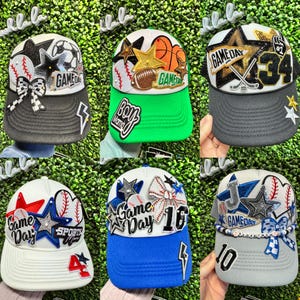 May include: Selection of baseball caps with sports-themed decorations. Each cap has a different colour scheme, with details such as stars, baseballs, and the words "Game Day" and numbers. The caps have curved peaks and adjustable fastenings.